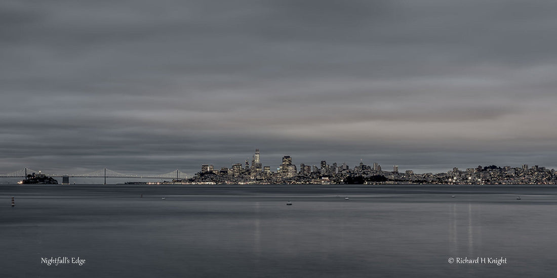 Nightfall’s Edge by Richard H Knight: San Francisco’s illuminated skyline, Alcatraz and the Bay Bridge across calm bay waters beneath a clouded evening sky
