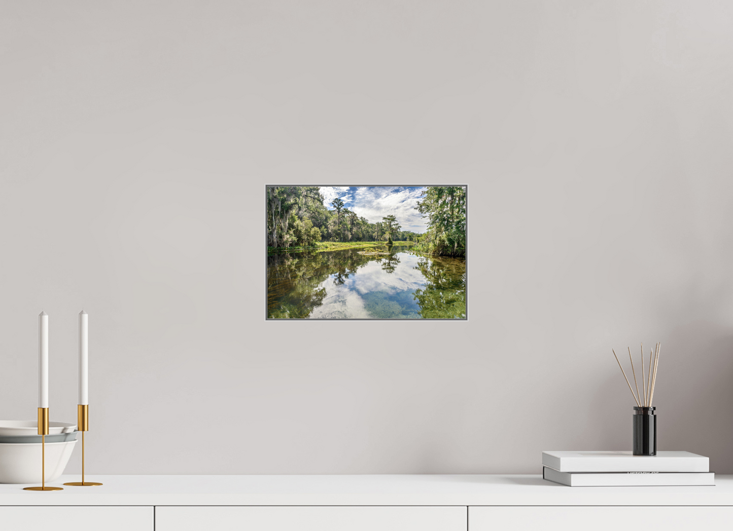 Scenic wall art of a lake with trees on a gray wall above a white surface.