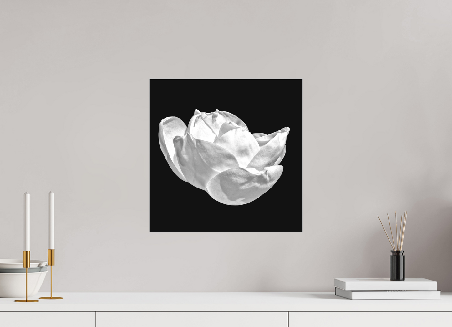 Framed black and white photograph of a flower on a wall above a white surface with decorative items.